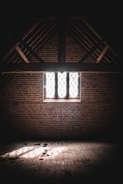 A dimly lit attic with exposed wooden beams and a brick wall. Sunlight filters through a latticed window, casting patterned shadows on the dusty wooden floor.