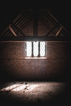 A dimly lit attic with exposed wooden beams and a brick wall. Sunlight filters through a latticed window, casting patterned shadows on the dusty wooden floor.