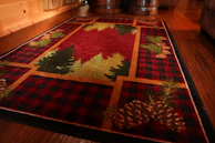 A rustic rectangular rug with a checkered pattern in red and black, featuring designs of green pine trees and brown pinecones, set on a wooden floor next to wooden barrels and furnishings.