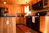 A cozy, warmly lit kitchen features rustic wooden cabinets and black appliances. The countertops are adorned with jars, a kitchen towel, and flowers in a metal container. Overhead lights and a small 'Welcome' sign add to the inviting atmosphere.