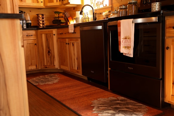 A warm and cozy kitchen with wooden cabinets and a dark countertop. The oven has a towel hanging that says 'Give Thanks'. Various kitchen items are neatly arranged, including a coffee maker and spice rack. The lighting gives a welcoming and rustic atmosphere.