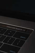 A close-up of a laptop keyboard featuring a prominent 'Unlock with Touch ID' button, surrounded by keys such as delete, return, and mathematical symbols on a dark background.