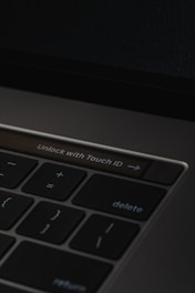 A close-up of a laptop keyboard featuring a prominent 'Unlock with Touch ID' button, surrounded by keys such as delete, return, and mathematical symbols on a dark background.