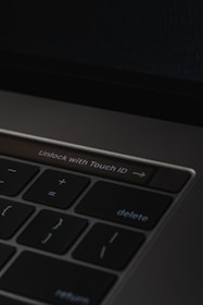 A close-up of a laptop keyboard featuring a prominent 'Unlock with Touch ID' button, surrounded by keys such as delete, return, and mathematical symbols on a dark background.