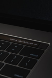 A close-up of a laptop keyboard featuring a prominent 'Unlock with Touch ID' button, surrounded by keys such as delete, return, and mathematical symbols on a dark background.