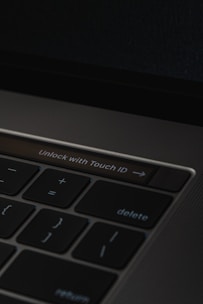 A close-up of a laptop keyboard featuring a prominent 'Unlock with Touch ID' button, surrounded by keys such as delete, return, and mathematical symbols on a dark background.