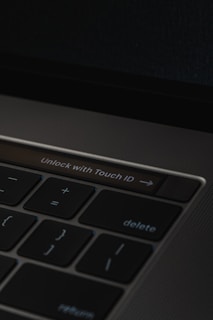 A close-up of a laptop keyboard featuring a prominent 'Unlock with Touch ID' button, surrounded by keys such as delete, return, and mathematical symbols on a dark background.