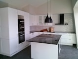 A modern kitchen with sleek white cabinets and a dark stone countertop. Two black pendant lights hang over a central island. There's a built-in oven and a white tile backsplash. A closed cardboard box is on the counter.