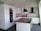 A modern kitchen with sleek white cabinets and a dark stone countertop. Two black pendant lights hang over a central island. There's a built-in oven and a white tile backsplash. A closed cardboard box is on the counter.
