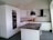 A modern kitchen with sleek white cabinets and a dark stone countertop. Two black pendant lights hang over a central island. There's a built-in oven and a white tile backsplash. A closed cardboard box is on the counter.