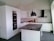 A modern kitchen with sleek white cabinets and a dark stone countertop. Two black pendant lights hang over a central island. There's a built-in oven and a white tile backsplash. A closed cardboard box is on the counter.