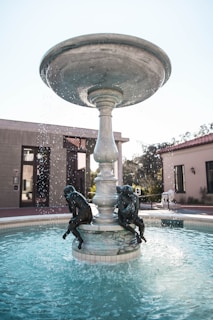 A large, tiered fountain with water gently cascading from the top bowl, adorned with bronze statues of children sitting around the base. The setting appears to be an outdoor plaza or courtyard, with modern buildings and some greenery visible in the background.