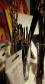 A variety of paintbrushes are placed inside a silver container on a desk. Surrounding the container, there are some art supplies, including packs of acrylic paint and possibly stretched canvases. Some dried branches can also be seen placed in a glass vase. The background consists of colorful, abstract artwork.