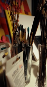 A variety of paintbrushes are placed inside a silver container on a desk. Surrounding the container, there are some art supplies, including packs of acrylic paint and possibly stretched canvases. Some dried branches can also be seen placed in a glass vase. The background consists of colorful, abstract artwork.