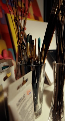 A variety of paintbrushes are placed inside a silver container on a desk. Surrounding the container, there are some art supplies, including packs of acrylic paint and possibly stretched canvases. Some dried branches can also be seen placed in a glass vase. The background consists of colorful, abstract artwork.