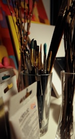 A variety of paintbrushes are placed inside a silver container on a desk. Surrounding the container, there are some art supplies, including packs of acrylic paint and possibly stretched canvases. Some dried branches can also be seen placed in a glass vase. The background consists of colorful, abstract artwork.