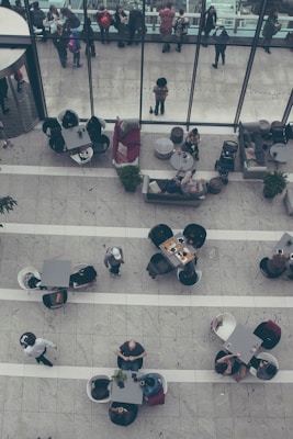 A top-down view of a spacious indoor area with multiple sets of tables and chairs. People are sitting at the tables, engaged in various activities such as talking, eating, and using electronic devices. Several groups are gathered near large windows at the far end. The area has a modern and minimalistic design with gray tiled flooring and sparse greenery.