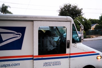 A United States Postal Service mail truck is parked on a residential street. The driver, partially visible through the window and dressed in a plaid shirt, is seated inside. The truck features the official USPS logo and is painted in white with red and blue stripes. Trees and a house are visible in the background.