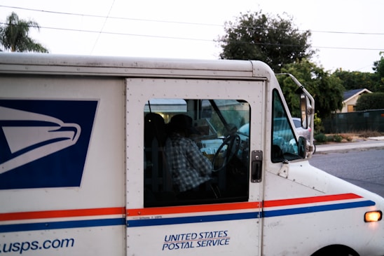 A United States Postal Service mail truck is parked on a residential street. The driver, partially visible through the window and dressed in a plaid shirt, is seated inside. The truck features the official USPS logo and is painted in white with red and blue stripes. Trees and a house are visible in the background.