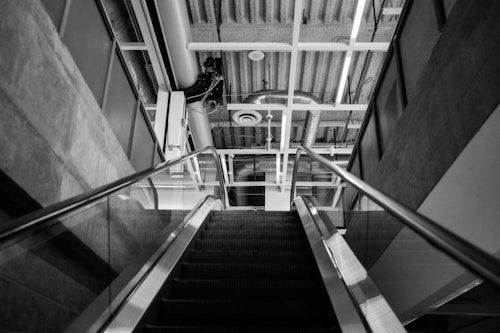 An escalator ascends towards a ceiling with exposed structural elements, including ducts and pipes. The architecture is modern and industrial, with clean lines and a monochrome palette. The view is upward, emphasizing the geometric patterns formed by the ceiling and escalator supports.