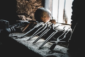 A collection of old, rusty tools is scattered on a dusty workbench in a dimly lit workshop. Sunlight filters through a nearby window, casting shadows on the rough stone walls, with various metal implements including tongs and pincers prominently displayed. A sphere or round object can be seen in the background, partially illuminated by the light.