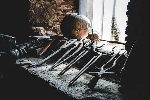 A collection of old, rusty tools is scattered on a dusty workbench in a dimly lit workshop. Sunlight filters through a nearby window, casting shadows on the rough stone walls, with various metal implements including tongs and pincers prominently displayed. A sphere or round object can be seen in the background, partially illuminated by the light.