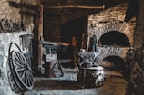 A rustic and dimly lit blacksmith's workshop with stone walls and a variety of blacksmithing tools. The space includes an anvil, a forge, several hammers, and tongs. Old wagon wheel and chains are also present, adding to the historical ambiance.