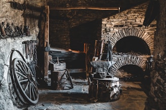 A rustic and dimly lit blacksmith's workshop with stone walls and a variety of blacksmithing tools. The space includes an anvil, a forge, several hammers, and tongs. Old wagon wheel and chains are also present, adding to the historical ambiance.