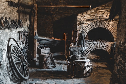 A rustic and dimly lit blacksmith's workshop with stone walls and a variety of blacksmithing tools. The space includes an anvil, a forge, several hammers, and tongs. Old wagon wheel and chains are also present, adding to the historical ambiance.