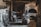 A rustic and dimly lit blacksmith's workshop with stone walls and a variety of blacksmithing tools. The space includes an anvil, a forge, several hammers, and tongs. Old wagon wheel and chains are also present, adding to the historical ambiance.