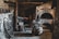 A rustic and dimly lit blacksmith's workshop with stone walls and a variety of blacksmithing tools. The space includes an anvil, a forge, several hammers, and tongs. Old wagon wheel and chains are also present, adding to the historical ambiance.