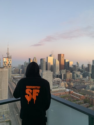 A person wearing a black hoodie stands on a balcony overlooking a cityscape. The hoodie has orange text and graphics on the back. The city has a mix of modern skyscrapers and older buildings, with a clear sky above.