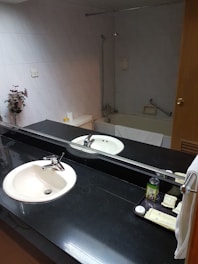 A bathroom featuring a black countertop with a white sink and chrome faucet, a large mirror reflecting the room, and various toiletries including a can and several packets on a tray. In the background, there is a bathtub with a showerhead and a towel draped over the edge. A floral arrangement is placed on the left side near a tiled wall.