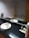 A bathroom featuring a black countertop with a white sink and chrome faucet, a large mirror reflecting the room, and various toiletries including a can and several packets on a tray. In the background, there is a bathtub with a showerhead and a towel draped over the edge. A floral arrangement is placed on the left side near a tiled wall.