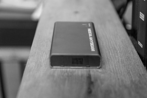 A compact electronic device with the label MEERA ANYBEAM resting on a wooden surface. The device appears to be sleek and rectangular, with a clean, modern design.