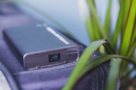A close-up of a compact black electronic device, resembling a power bank or modem, placed on a dark fabric surface. To the right are green leaves from a plant, partially in focus, adding a natural element to the composition.