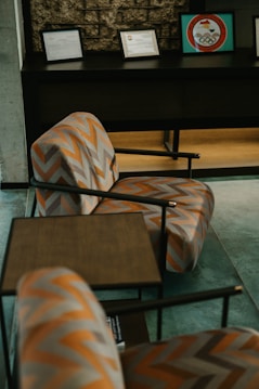 A room with a patterned armchair featuring a zigzag orange and gray design. A small wooden table is placed next to the chair. In the background, various framed certificates or awards are displayed on a dark console table against a textured brick wall. The overall setting suggests a professional or lounge space.