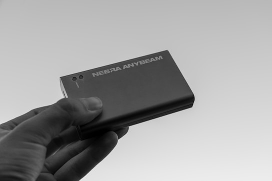 A hand is holding a compact, rectangular device labeled 'NEBRA ANYBEAM'. The device appears sleek and modern with a metallic finish.