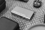 A small electronic device labeled with the words 'NEBRA ANYBEAM' lies on a patterned carpet. Surrounding it are various other electronic accessories including a power adapter.
