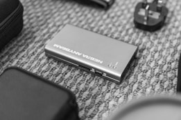 A small electronic device labeled with the words 'NEBRA ANYBEAM' lies on a patterned carpet. Surrounding it are various other electronic accessories including a power adapter.