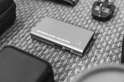 A small electronic device labeled with the words 'NEBRA ANYBEAM' lies on a patterned carpet. Surrounding it are various other electronic accessories including a power adapter.