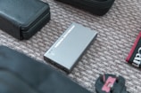 A NEBULA ANYBEAM device, possibly a mini projector, is placed on a textured surface alongside a black leather pouch and other electronic accessories, including a power adapter. The composition suggests a collection of travel or tech gear.