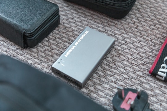 A NEBULA ANYBEAM device, possibly a mini projector, is placed on a textured surface alongside a black leather pouch and other electronic accessories, including a power adapter. The composition suggests a collection of travel or tech gear.