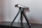 A compact, silver and black electronic device is mounted on a small tripod and placed on a dark wooden surface. The device appears to have various ports and buttons, suggesting functionality such as a camera or projector. The tripod's legs are extended, providing stability.
