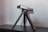 A compact, silver and black electronic device is mounted on a small tripod and placed on a dark wooden surface. The device appears to have various ports and buttons, suggesting functionality such as a camera or projector. The tripod's legs are extended, providing stability.
