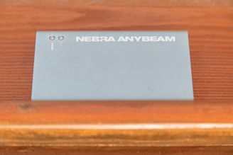 A device labeled Nebra Anybeam is placed on a wooden surface. The device is rectangular and metallic, with two ports labeled HDMI on one end. The surface it rests on is a warm-toned wood with horizontal lines.