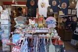 A vibrant storefront displaying a variety of traditional Japanese items including Maneki-neko statues, decorative fans, t-shirts with Japanese motifs, and an assortment of small souvenirs. The arrangement of the items creates an inviting and colorful atmosphere.