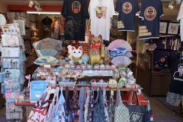 A vibrant storefront displaying a variety of traditional Japanese items including Maneki-neko statues, decorative fans, t-shirts with Japanese motifs, and an assortment of small souvenirs. The arrangement of the items creates an inviting and colorful atmosphere.