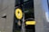 A decorative sign for a cafebar called 'GINZA 328' with a modern design is displayed against a backdrop of a large, reflective building with stone and glass elements. The sign features a bright yellow circular and rectangular design with artistic metalwork, depicting a steaming mug that suggests a welcoming environment.