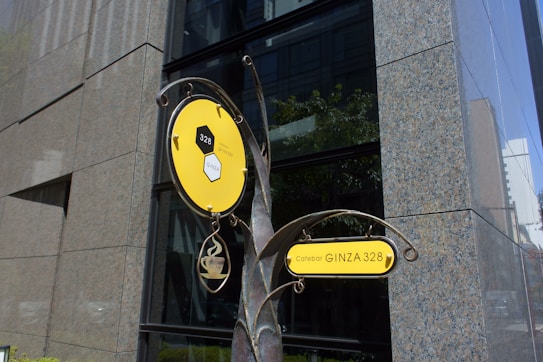 A decorative sign for a cafebar called 'GINZA 328' with a modern design is displayed against a backdrop of a large, reflective building with stone and glass elements. The sign features a bright yellow circular and rectangular design with artistic metalwork, depicting a steaming mug that suggests a welcoming environment.